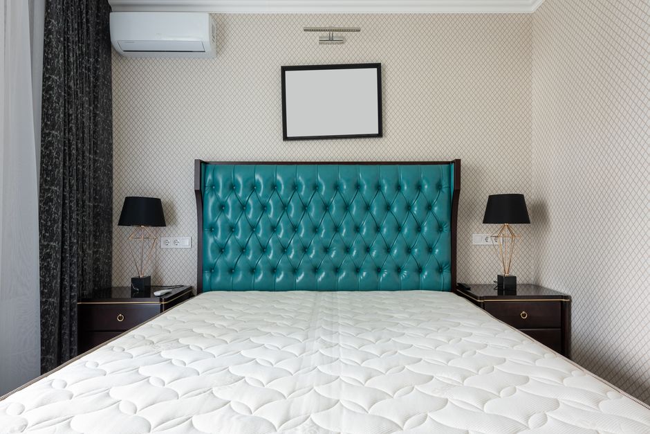 A person is making a bed with white bed linen in a bedroom featuring a wooden headboard, a bedside table with a brass lamp, and beige curtains. The individual is smoothing the duvet or sheet, which ap