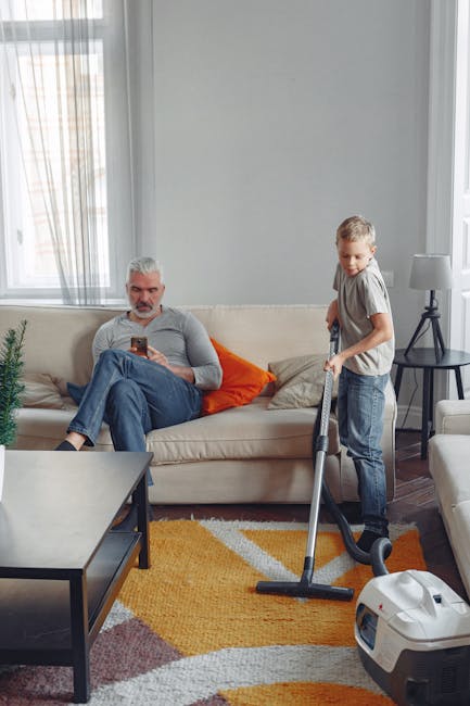 A living room with a light grey wall and large window covered by sheer curtains allowing natural light. A beige sofa with orange and beige cushions is positioned against the wall, and an older man wit