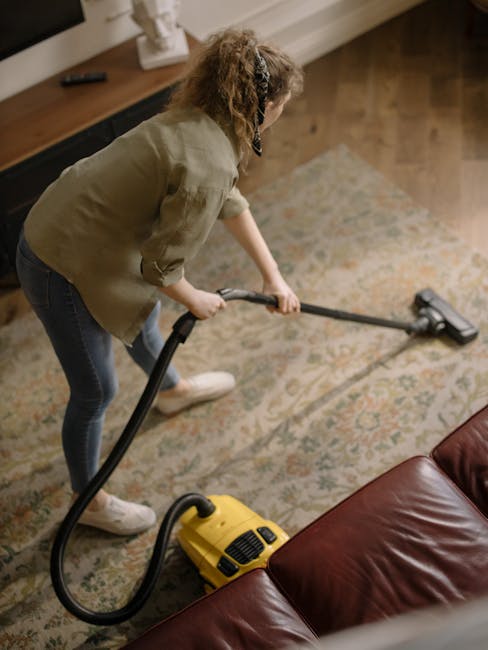 A person using a yellow portable industrial fan for surface cleaning or drying on a patterned, plush carpet in an indoor setting. The photo shows the top view of the person's hand pressing a control b