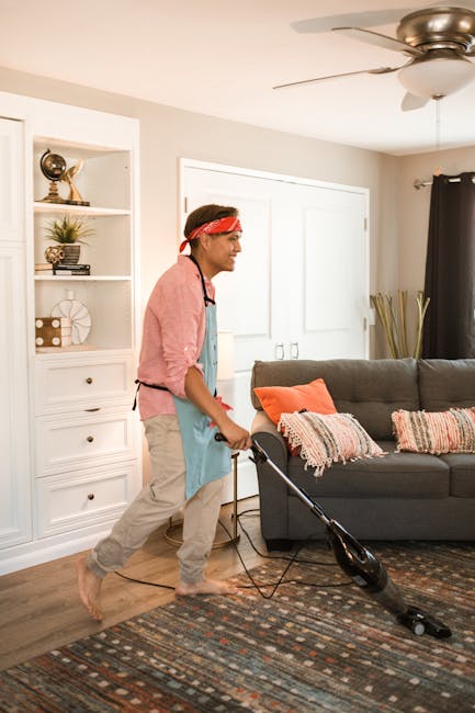 A close-up image of a traditional upright vacuum cleaner with a black and grey body, actively cleaning a patterned area rug in a living room. The vacuum's head rests flat on the floor, with the handle